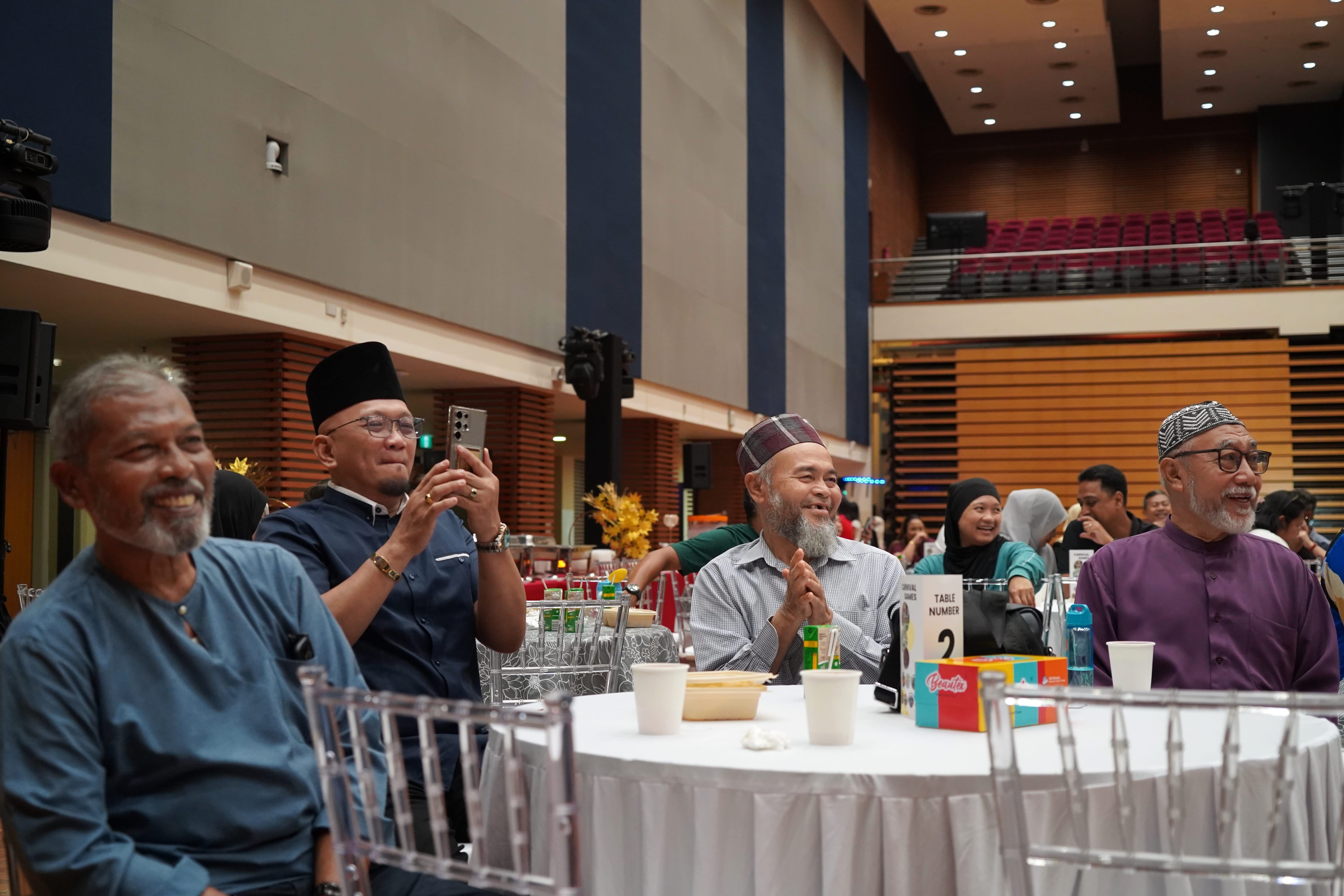 Several attendees seated around a banquet table smile, clap and watch the stage performance.